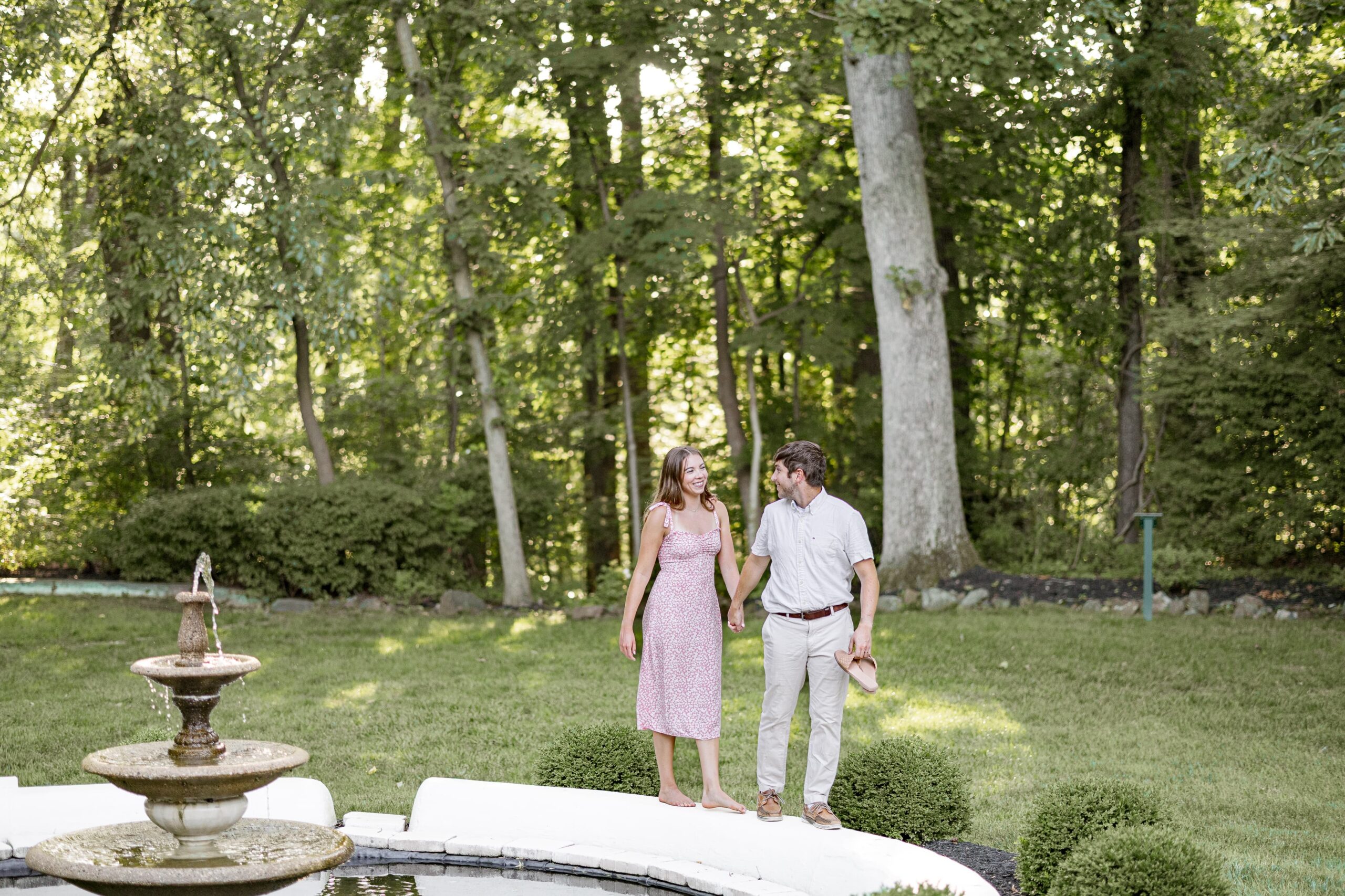Engaged man and woman smiling at each other as they walk along the edge of a water fountain. They are surrounded by greenery and trees.