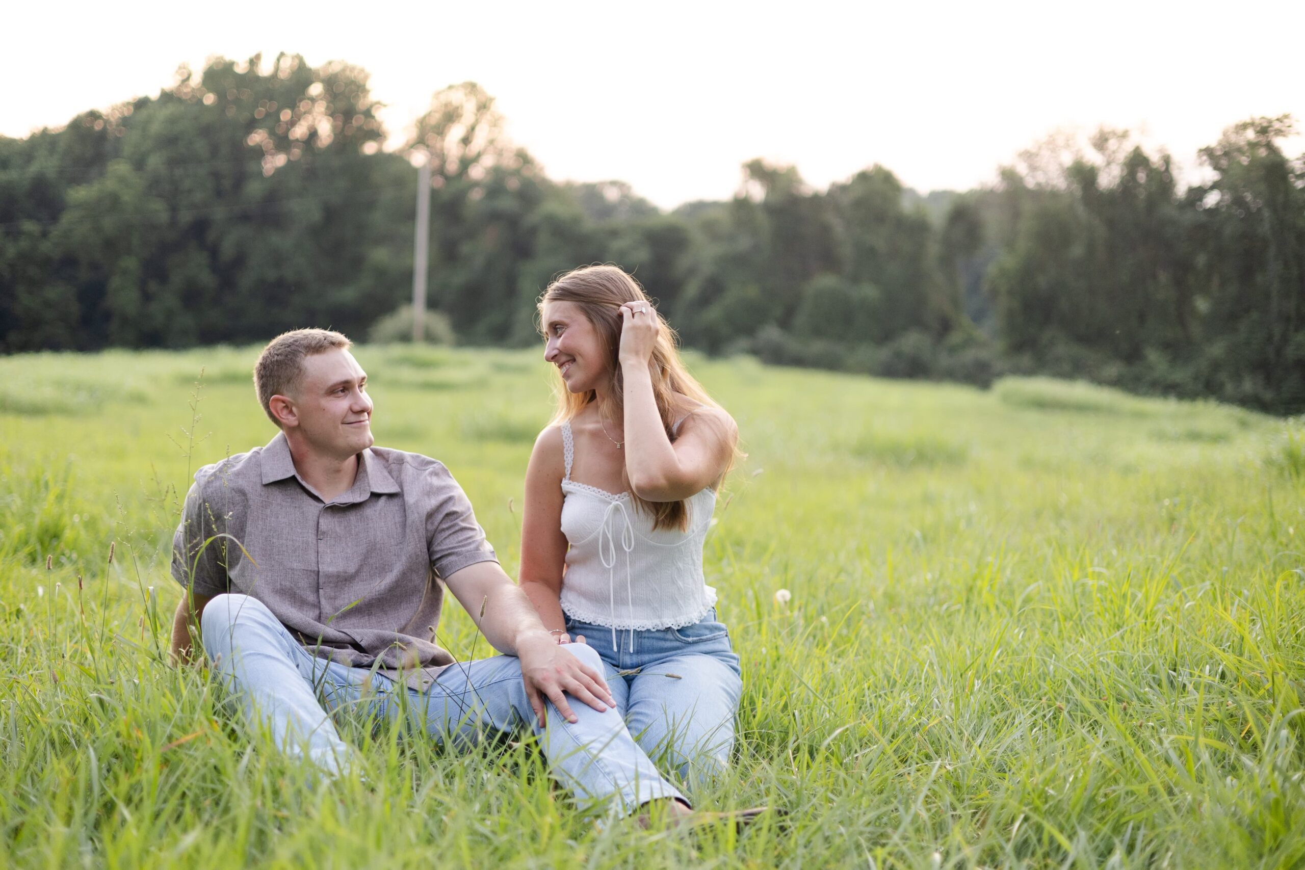 Man and woman sit in a grassy field. Both smiling at each other as the woman adjusts her hair with her hands. They are wearing jeans and neutral colored shirts