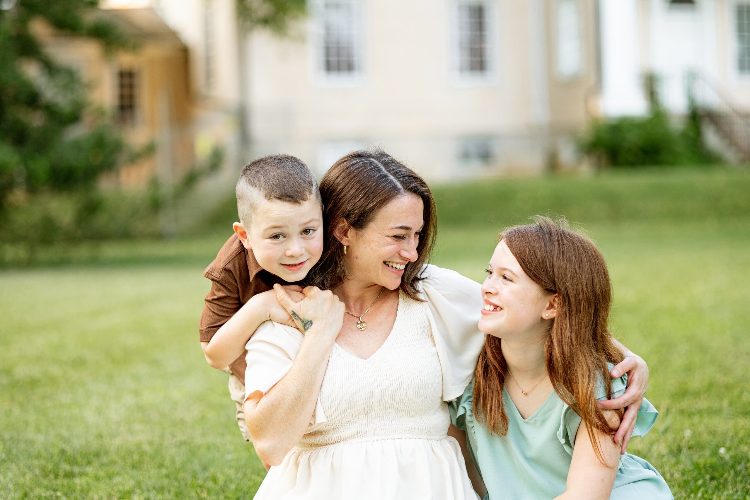 Mom and daughter smile at each other while son smiles at the camera. They are sitting in a grassy field in front of a historic mansion.