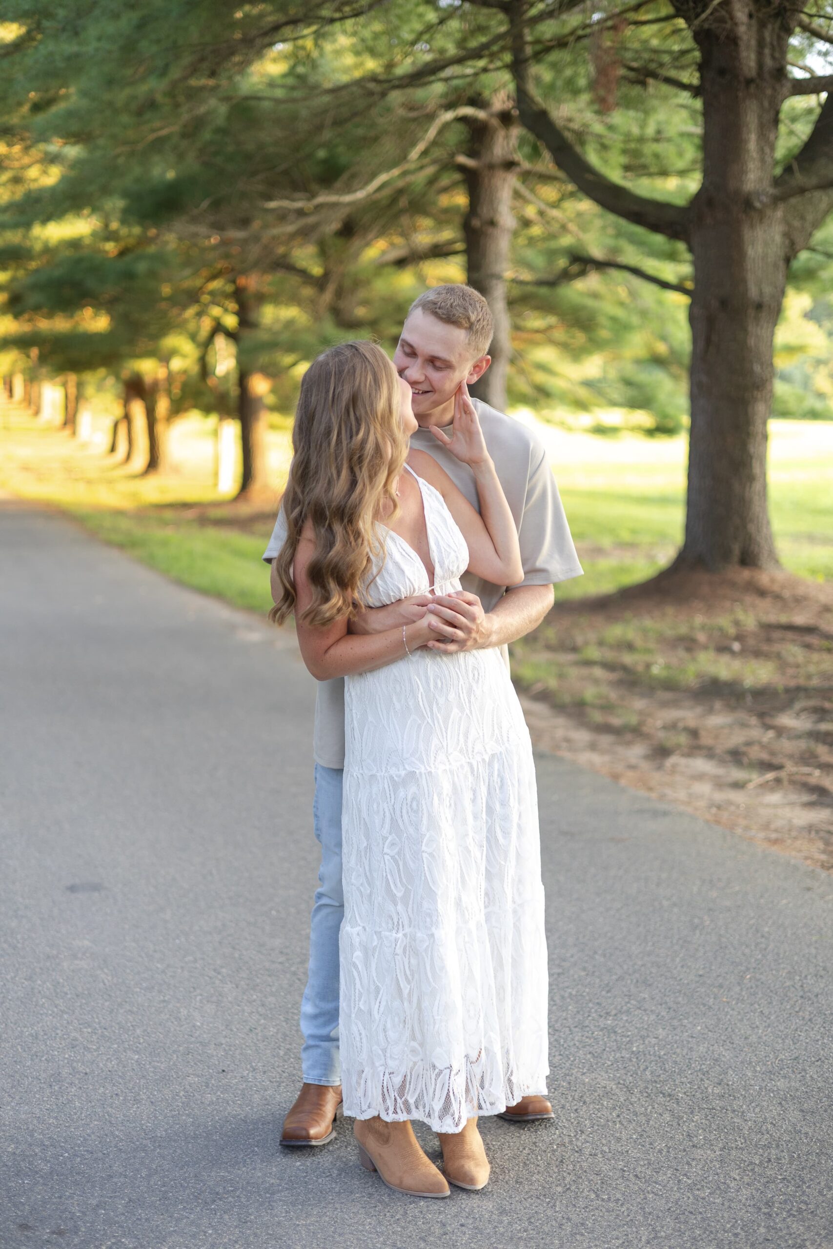 Woman in white dress and man in jeans and neutral shirt hug each other close and lean in for a kiss. They are smiling and standing on a gravel path.