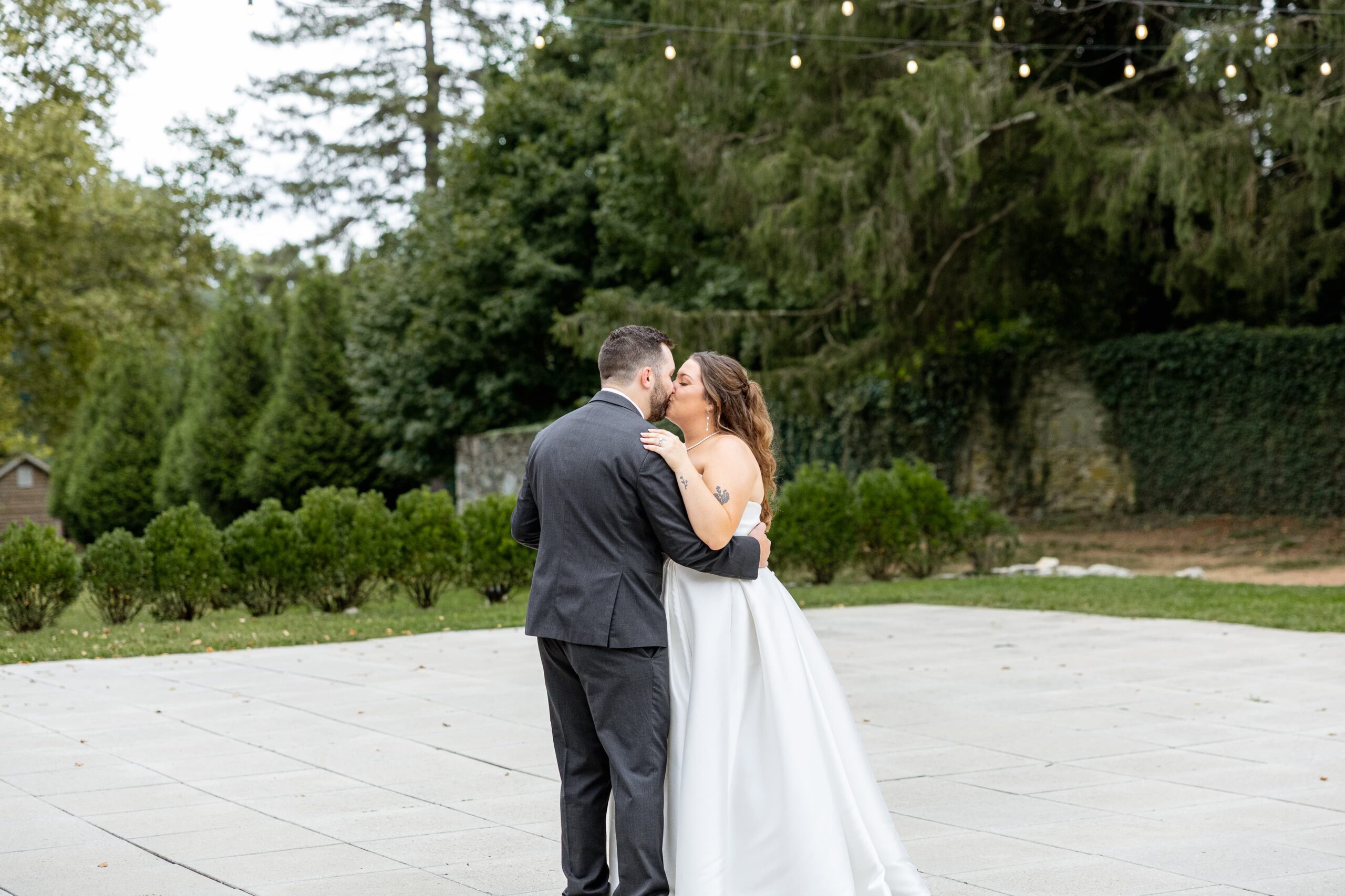 bride and groom sharing first dance on dance floor under outdoor string of lights and green trees.